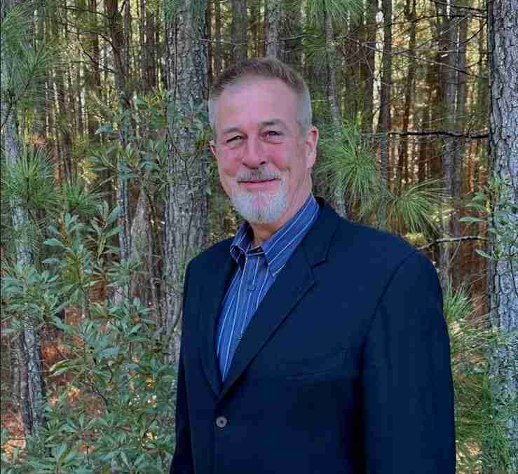A white man in a blue suit stands in front of a pine forest. He is smiling at the camera.