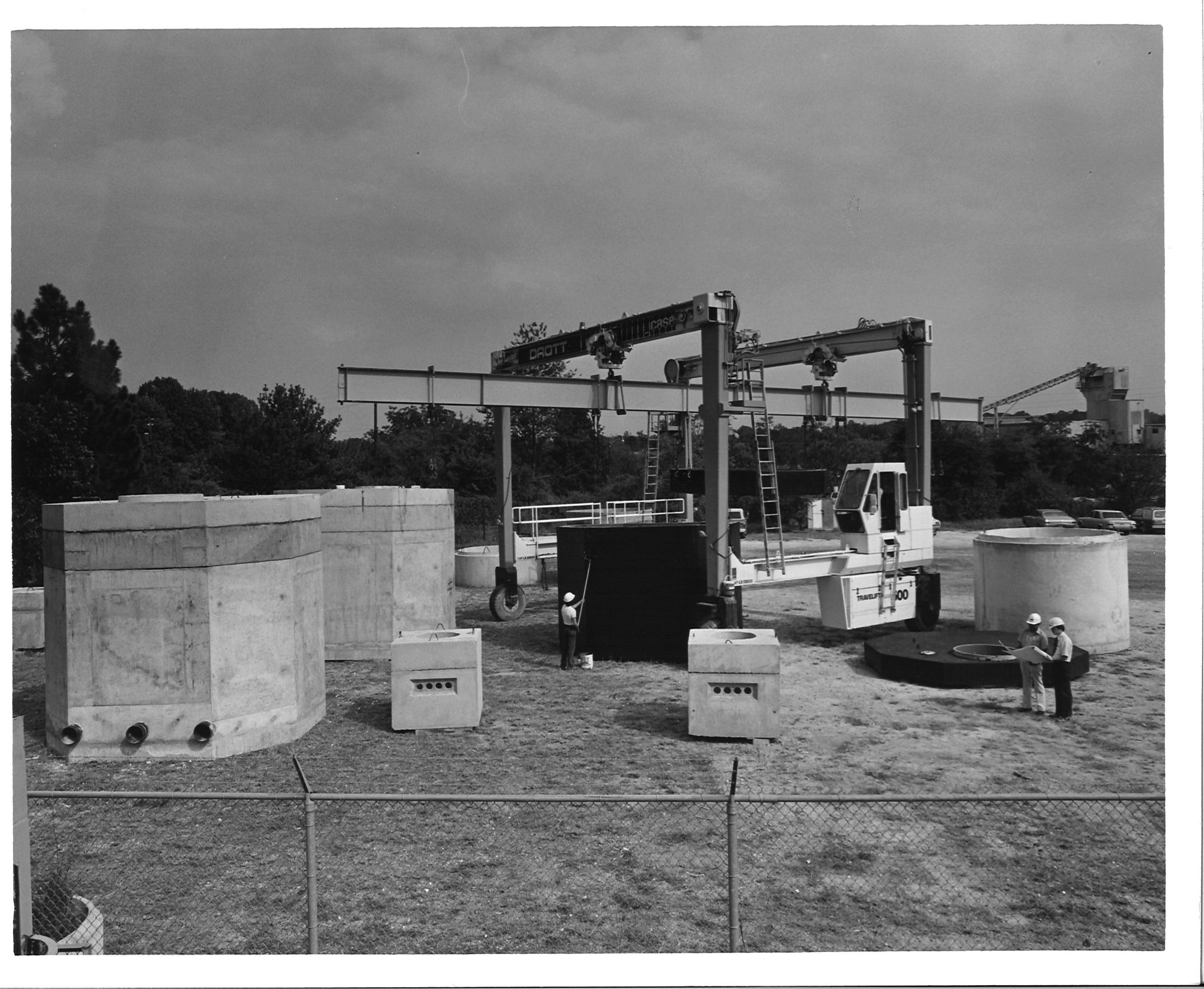 Construction site with precast concrete elements in progress. A few workers can be seen working on the foundation of a building with heavy machinery in the background.