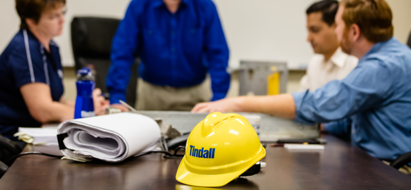 Several Tindall employees sit at a conference table. Many papers can be seen on the table. A yellow Tindall hard hat is centered and in focus in the photograph.